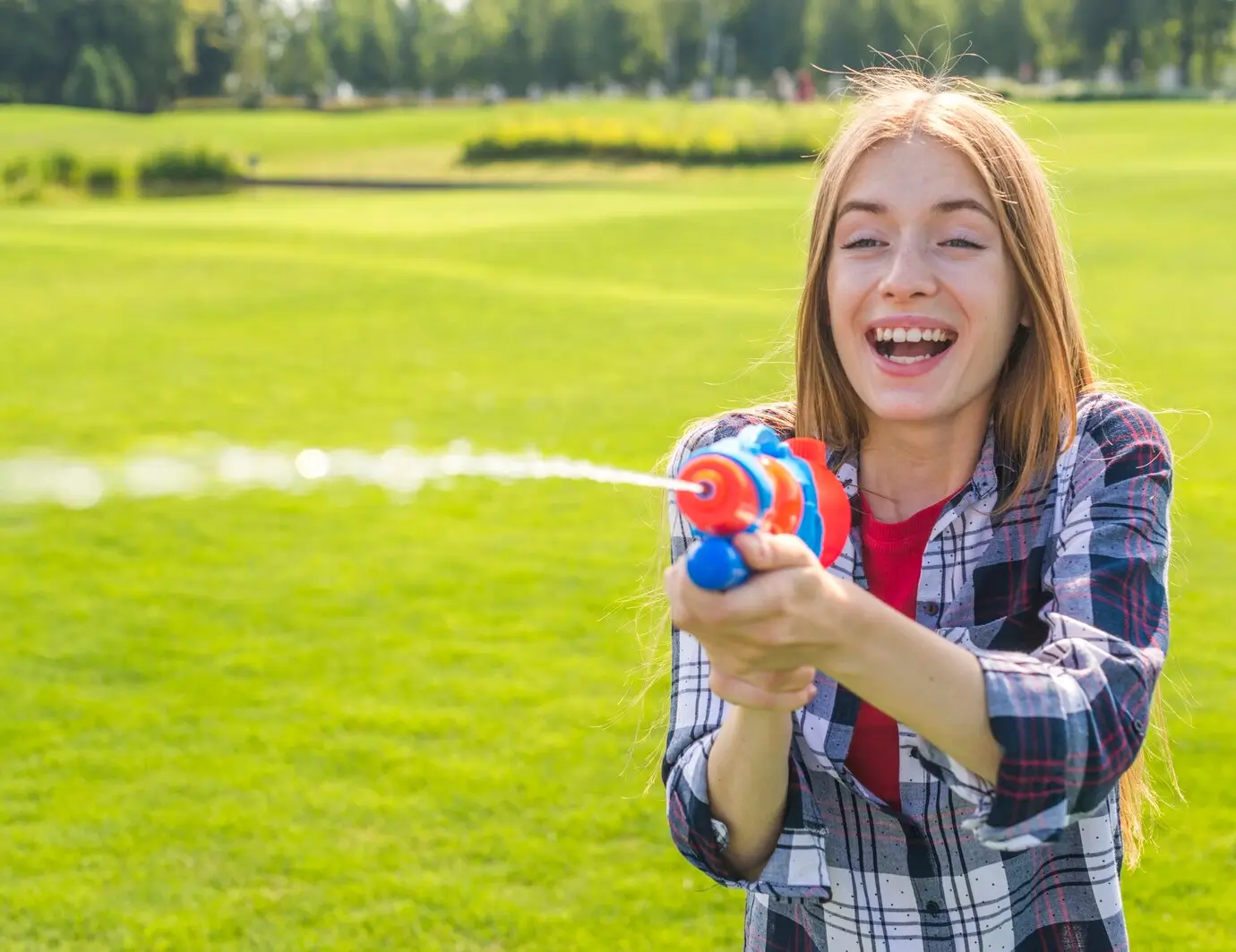 A happy woman with long red hair smiles at the camera while playfully spraying a water gun across a vibrant, green lawn of Texas Tuff artificial grass on a sunny day.
