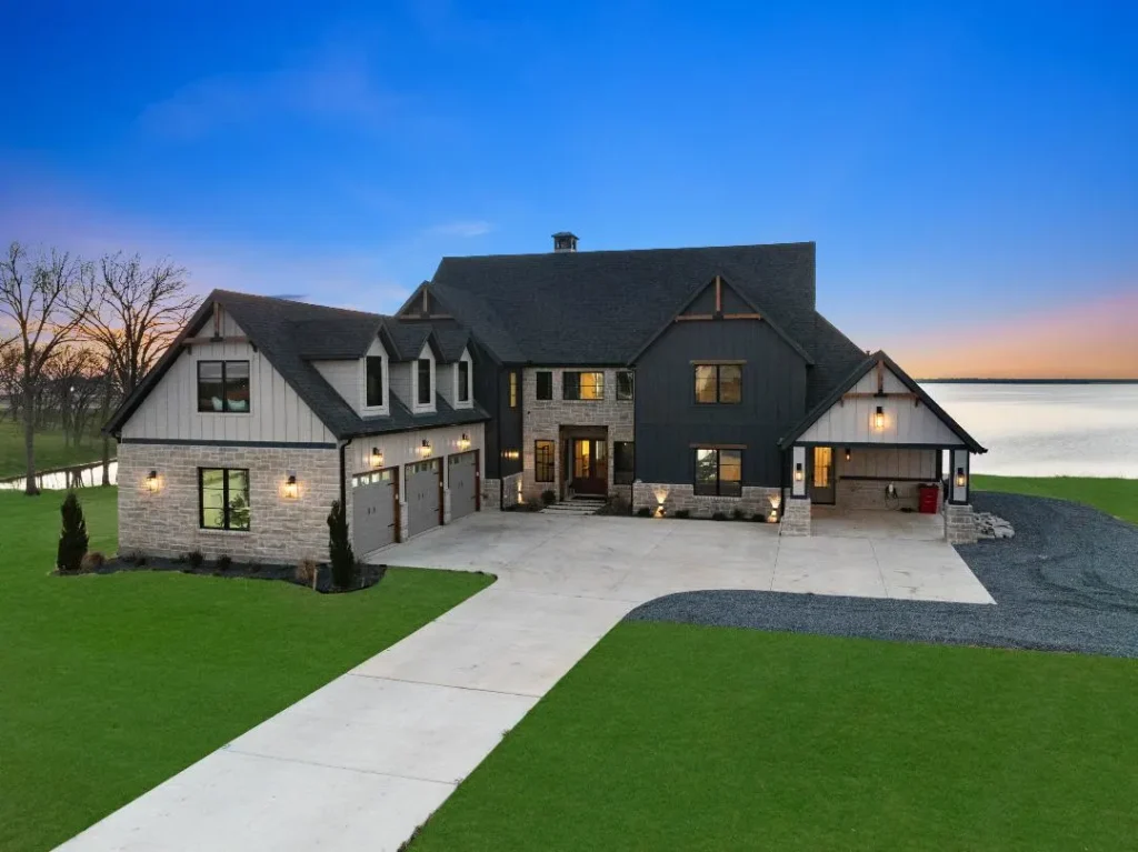 A breathtaking, newly constructed modern farmhouse-style mansion at twilight near a serene lake, featuring a sprawling green lawn, potentially showcasing a Turf Installation in Dallas. The grand home boasts a multi-car garage, a mix of stone and dark siding, and warmly lit windows, suggesting luxurious lakeside living with the potential for a professionally installed artificial turf lawn, similar to high-quality Turf Installation services available in Dallas.