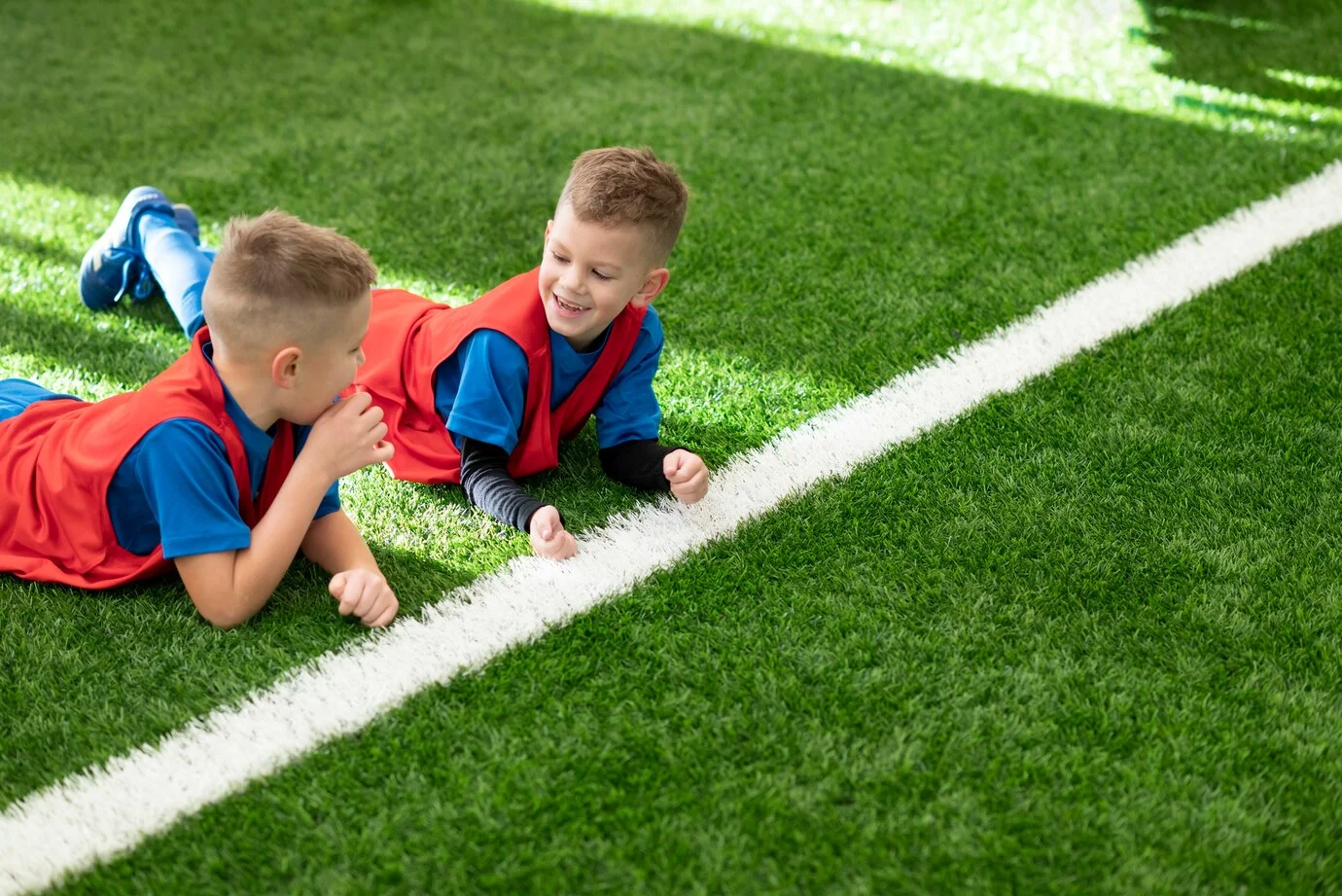 Two young boys in athletic wear are playfully interacting while lying on a vibrant green playground turf installation in Dallas. The artificial grass provides a safe and soft surface for children's play, indicated by the white line marking on the turf. This durable and low-maintenance playground turf is an ideal surfacing solution for parks, schools, and home playgrounds in the Dallas area, ensuring a clean and enjoyable play environment.